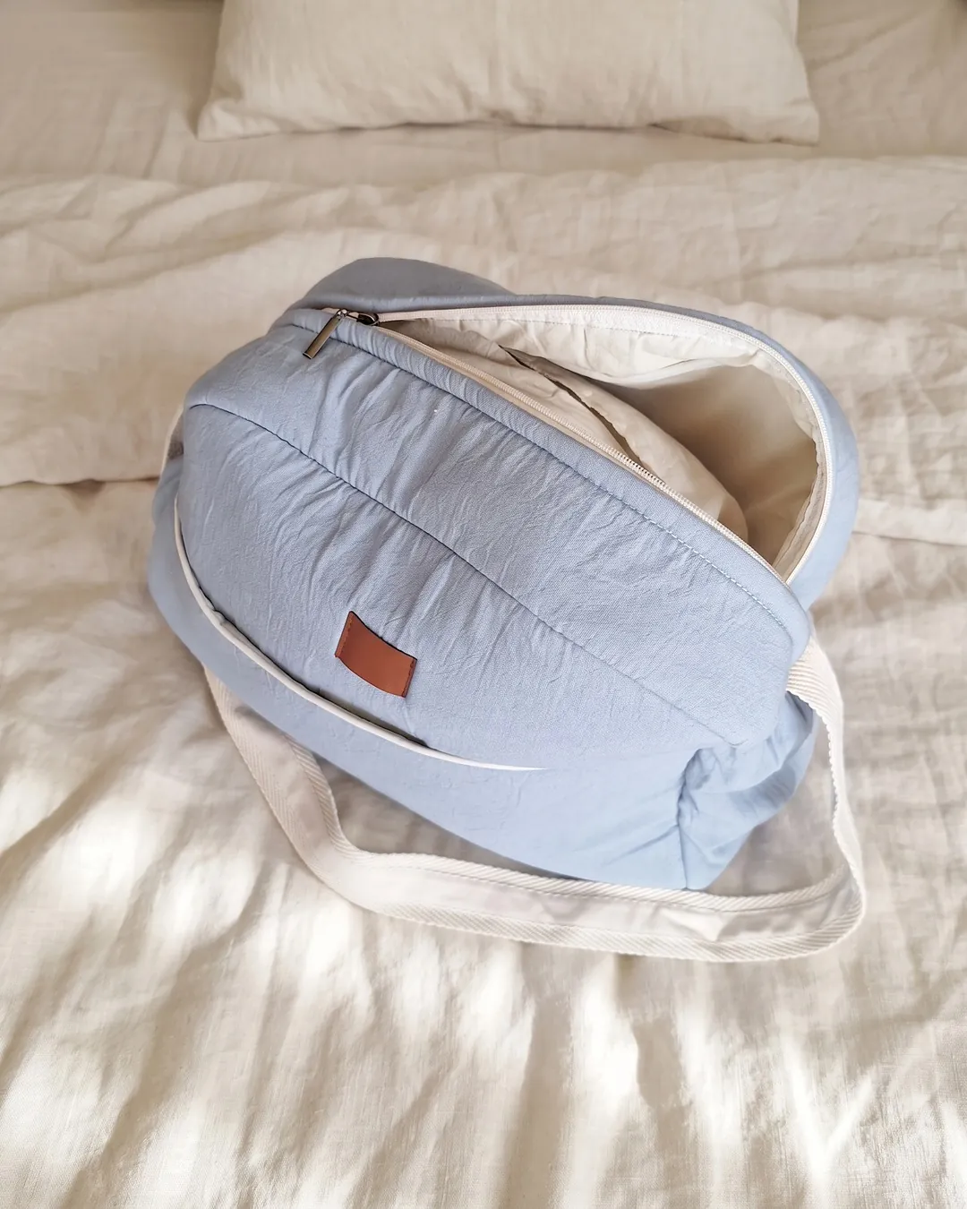 Bolso maternal celeste viejo con el cierre de arriba abierto, mostrando el interior forrado en crudo, sobre la cama.