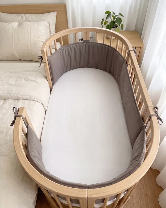 Cuna colecho ovalada de madera con un lado abierto junto a la cama de los padres, con chichonera acolchada gris topo.