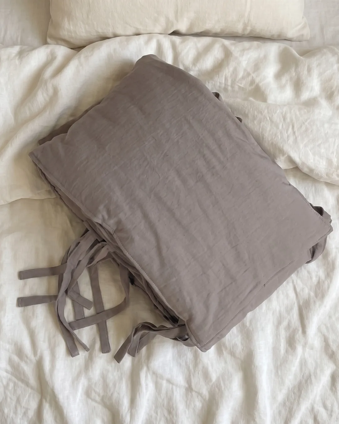 Chichonera gris topo doblada, con sus tiras para atar, sobre una cama con acolchado de lino crema.