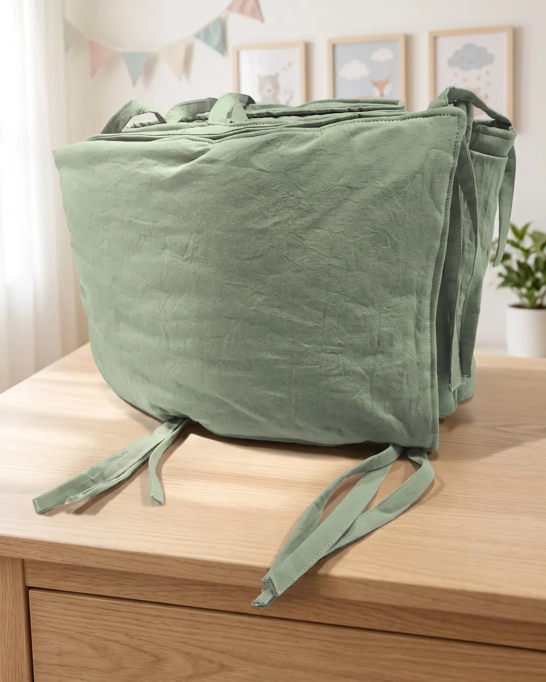 Chichonera verde seco doblada, con sus tiras para atar, sobre una cómoda de madera en un cuarto de bebé.