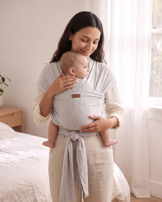 Mamá llevando a su bebé en un fular portabebé elástico de algodón gris melange, de pie junto a una ventana.