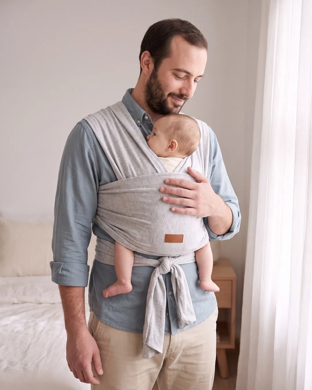 Papá con camisa celeste llevando a su bebé en un fular elástico de algodón gris melange atado adelante.