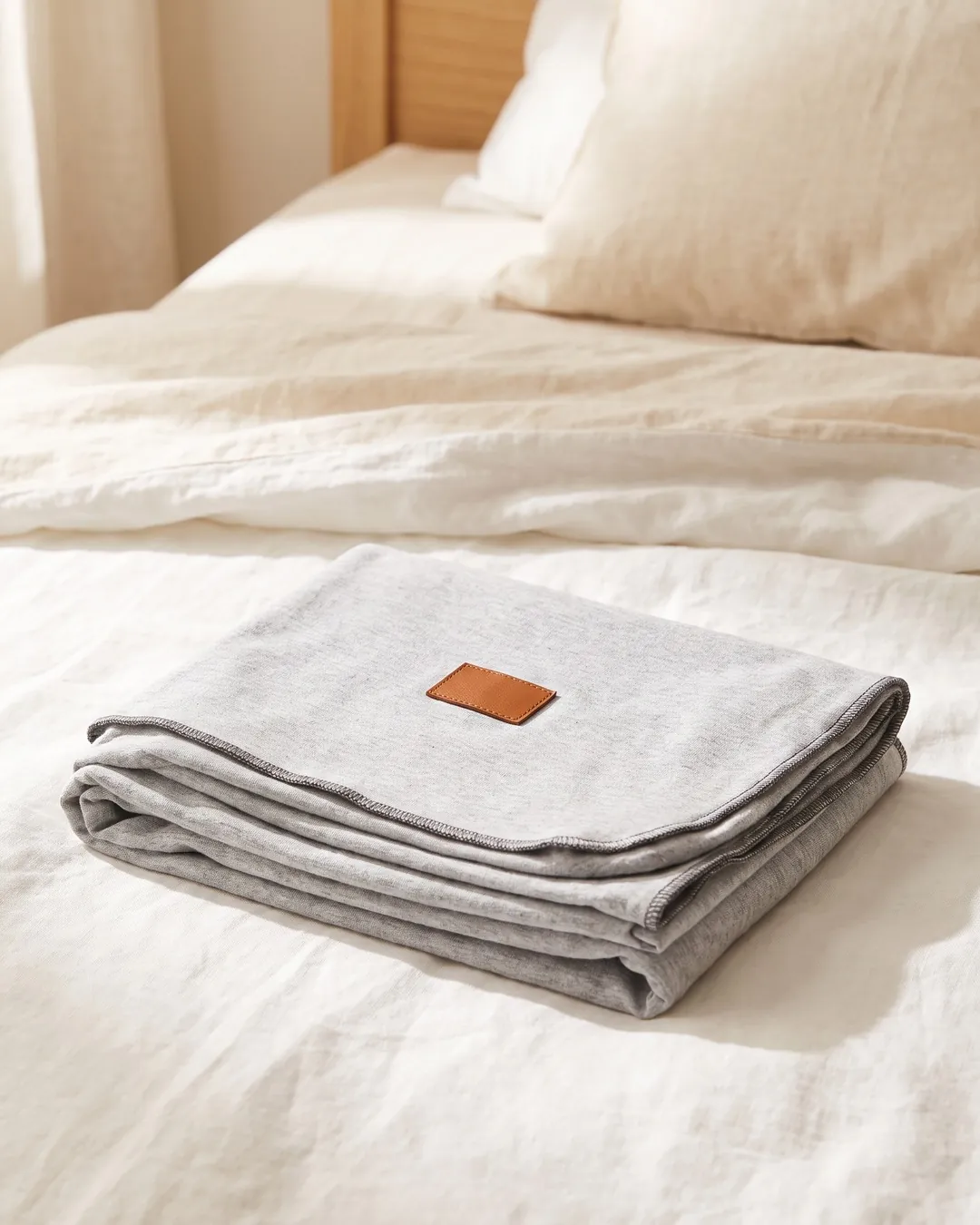 Fular elástico de algodón gris melange doblado sobre una cama, con un parche de cuero marrón.
